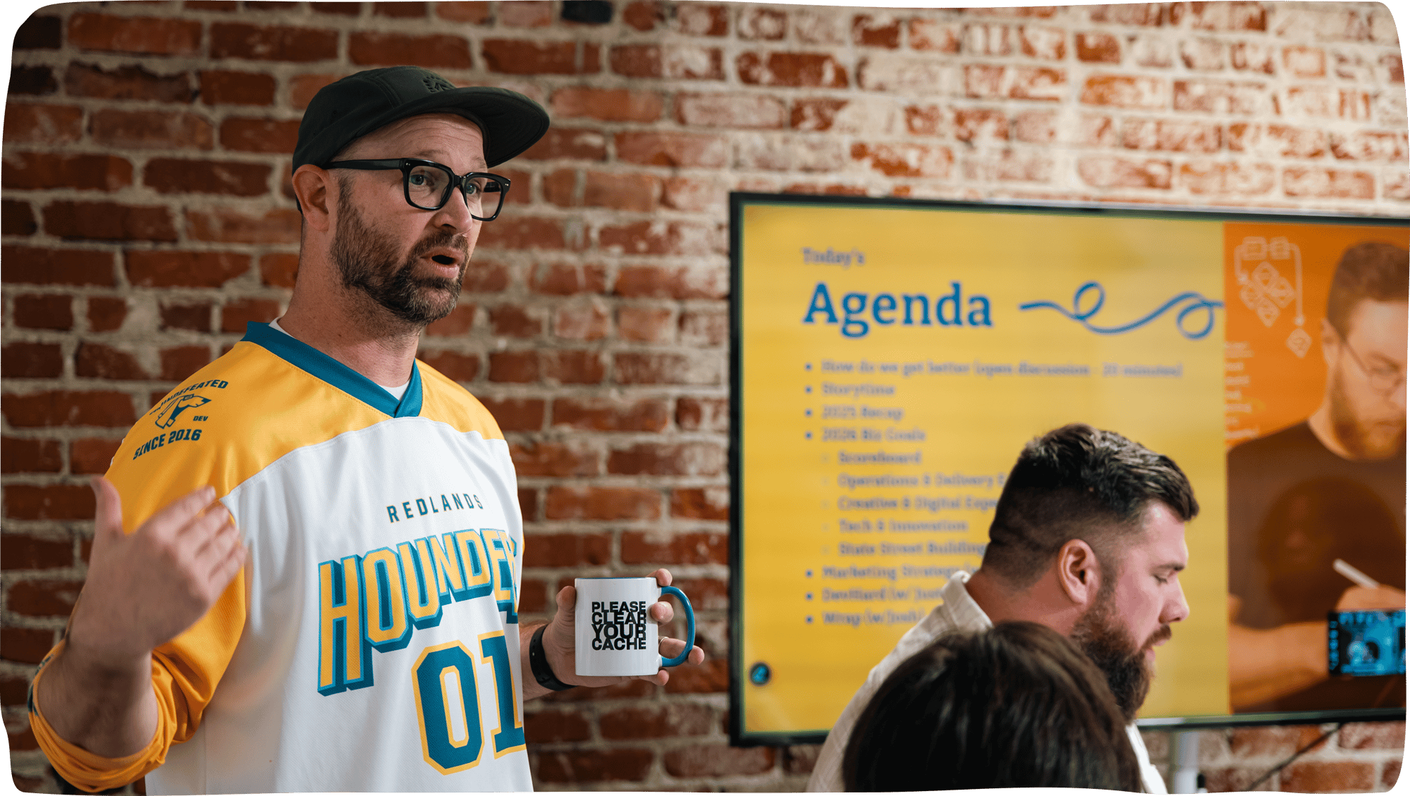 Hounder CEO standing with a coffee mug in front of a TV showing a slide presentation.